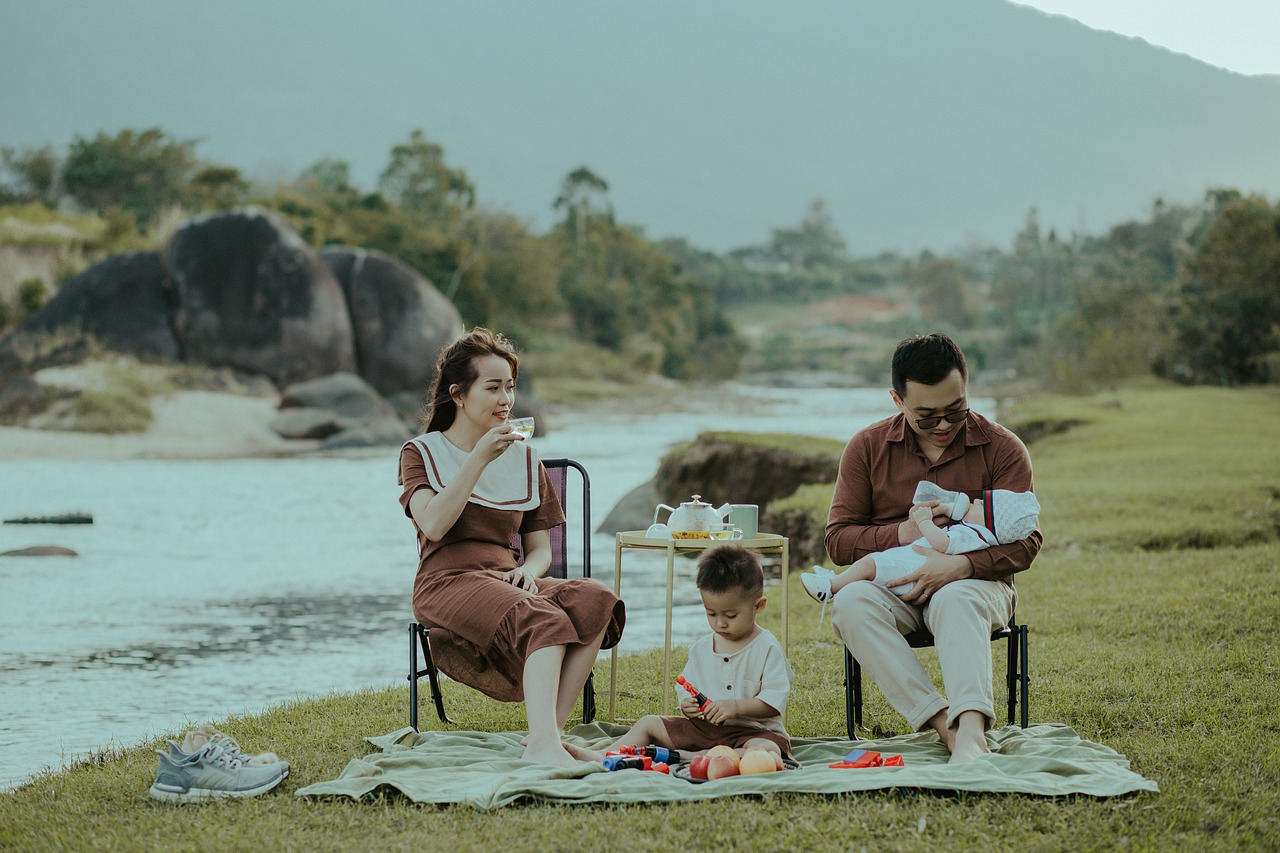 A family enjoying a sunny day, sitting on the grass by a serene river, surrounded by nature's beauty.